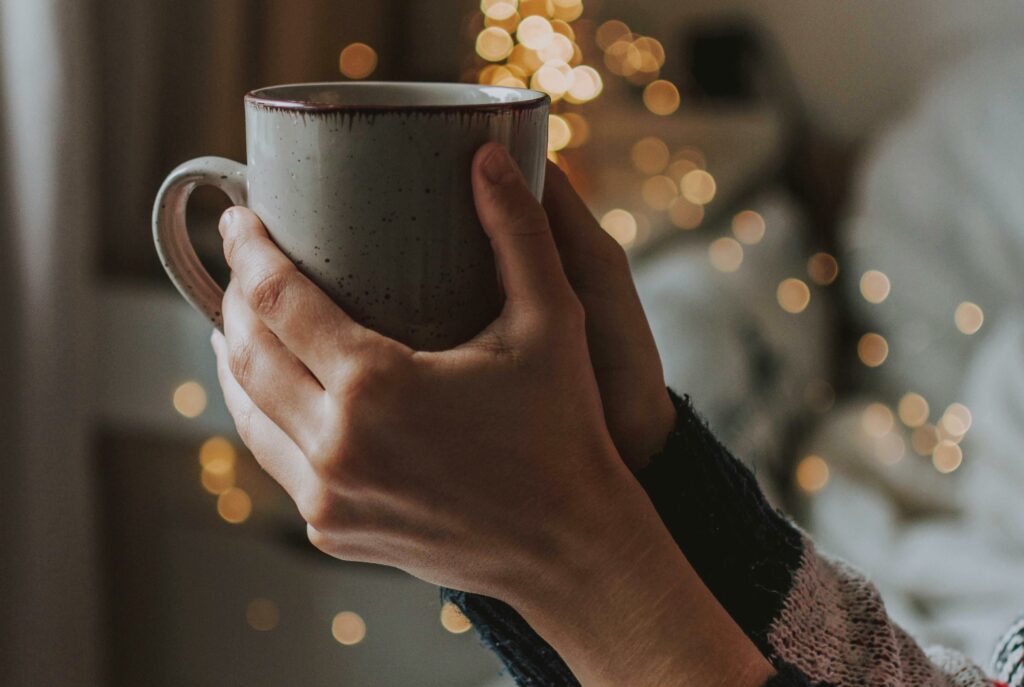 Two hands holding a coffee mug up with blurred twinkle lights in the distance.